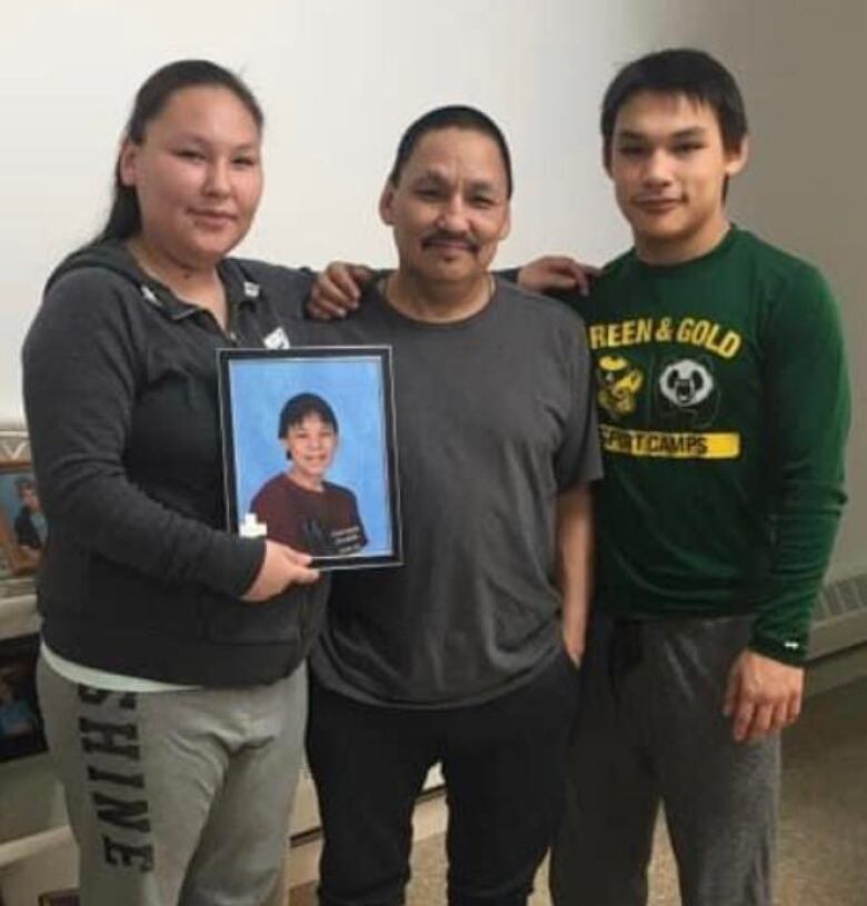 A woman and two men stand side by side, smiling for the camera, their arms around each other's shoulders. The woman his holding a framed photograph of a young man. 