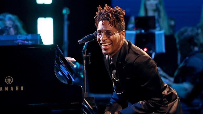 A man sits at a piano, leaning over and smiling towards the camera. 