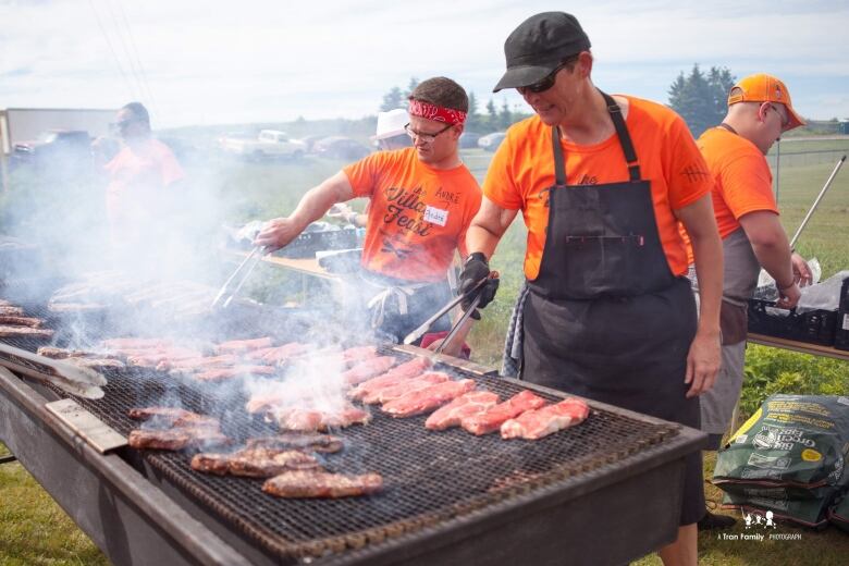 A man and woman wearing bright orange t-shirts cooking different meats on a large grill.