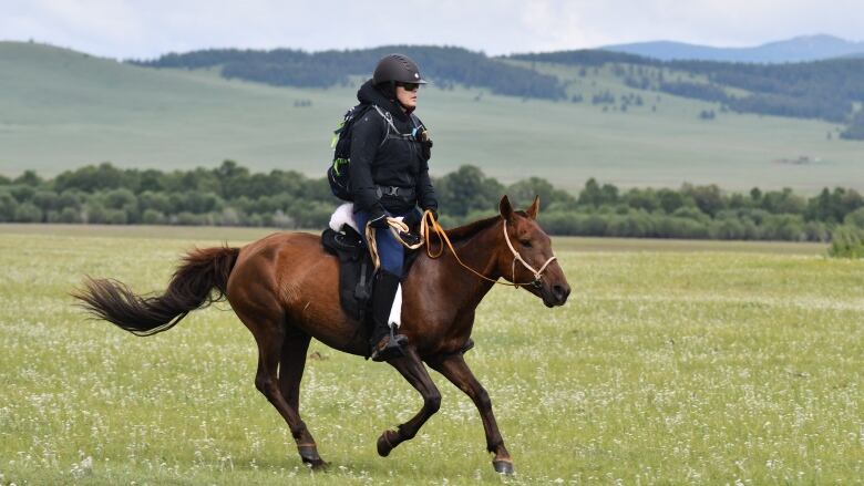 A woman wearing black gear rides a brown horse through the Mongolian countryside.