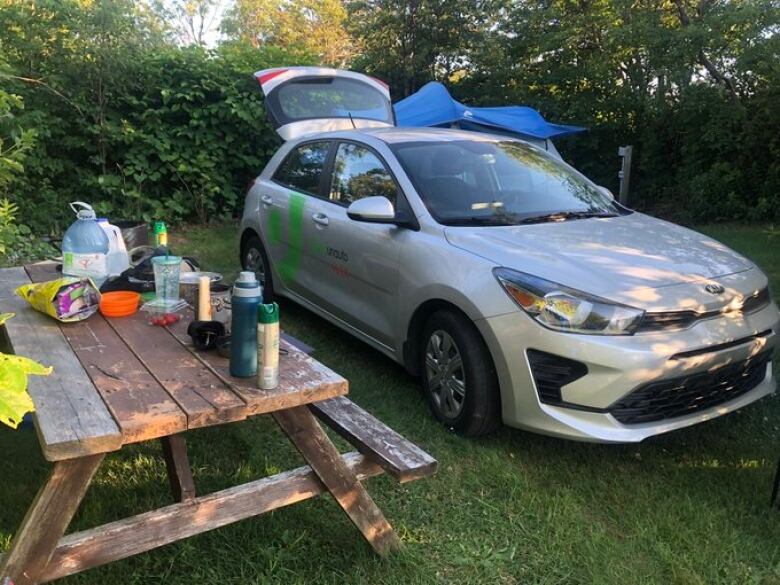 A car-sharing vehicle is parked in front of a tent at a campsite. A picnic table is covered in food and drinks in the foreground.