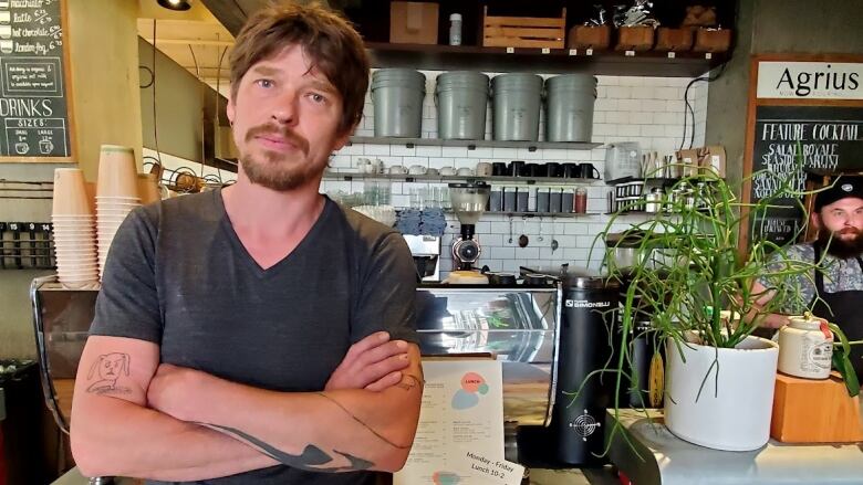 Cliff Leir folds his arms in front of a coffee shop counter. He is a white man with a moustache and goatee, wearing a dark grey shirt.