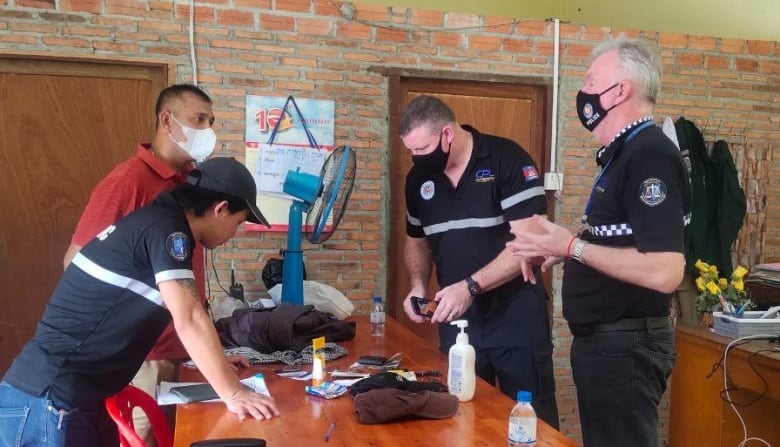 A group of masked men look at items on a table. Many of them are wearing police uniforms. A table fan and a sanitizer are visible on the table.