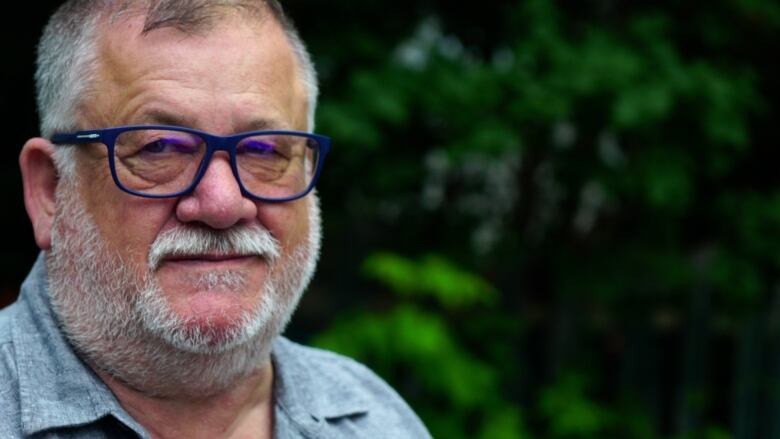 A man in his sixties with blue-framed glasses and stubble stares off. Dark green trees behind him. 