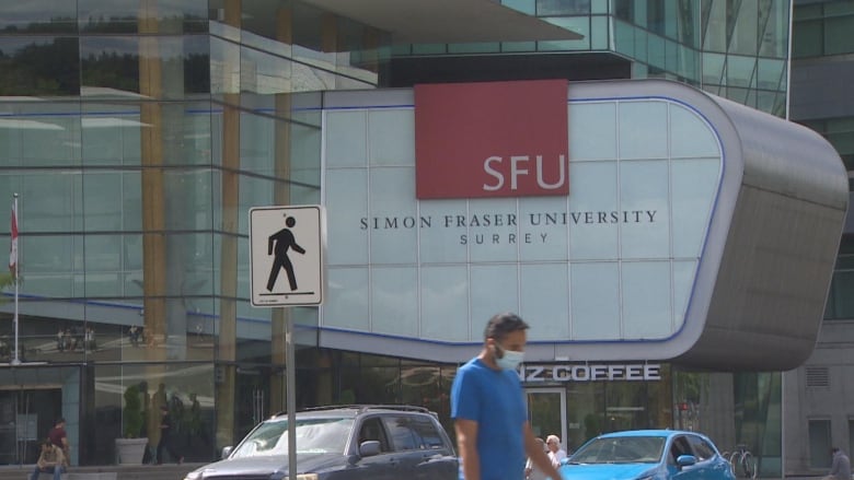 A person walks across the street near a campus building with the letters 'SFU' on it.