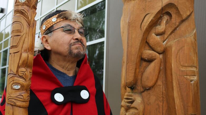 A man carrying a miniature totem pole stands next to a larger totem pole outside a building.