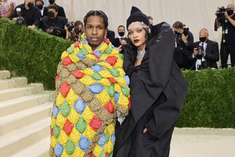 A man wrapped in a brightly coloured quilt stands closely with a woman wearing a billowing black dress and expensive jewelry. Behind them is a row of photographers. 