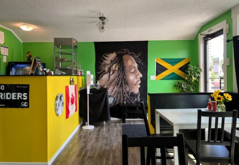 A small restaurant has a bright yellow till perpendicular to a green wall  the colours of the Jamaican flag. Canadian and Jamaican flags hang on the wall, as well as a large image of Bob Marley and a lion.