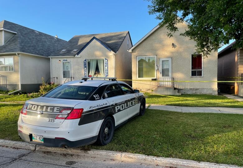 A Winnipeg police cruiser is parked on a boulevard in front of a single-storey house, which is surrounded in yellow police tape.