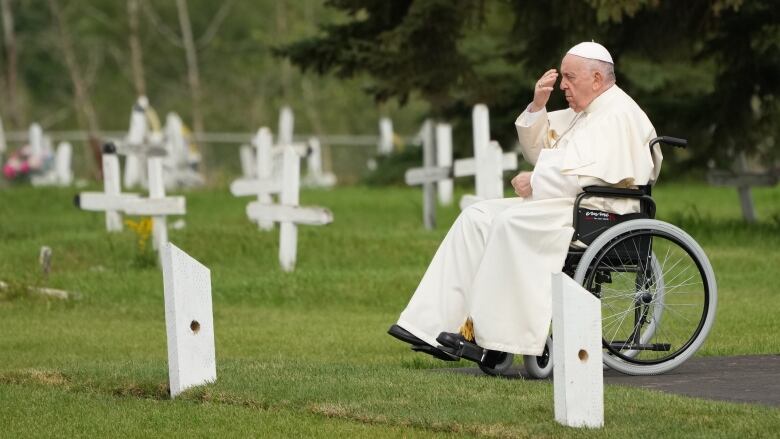 Pope Francis sits in his wheelchair at a gravesite at the Ermineskin Cree Nation Cemetery, raising his hand in a prayer motion.