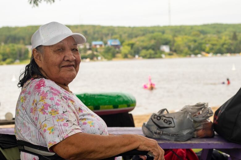 A woman in a patterned shirt and a white cap sits in a chair on the beach.