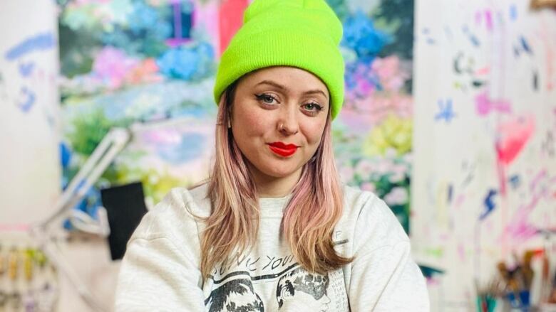 Photo of the artist Shanna Van Maurik, standing in her studio. She is a young white woman with rose-gold hair. She wears a neon green toque and a Sonic Youth sweatshirt. She smirks and the camera and crosses her arms in front of her. A colourful painting can be seen hanging on the wall behind her.