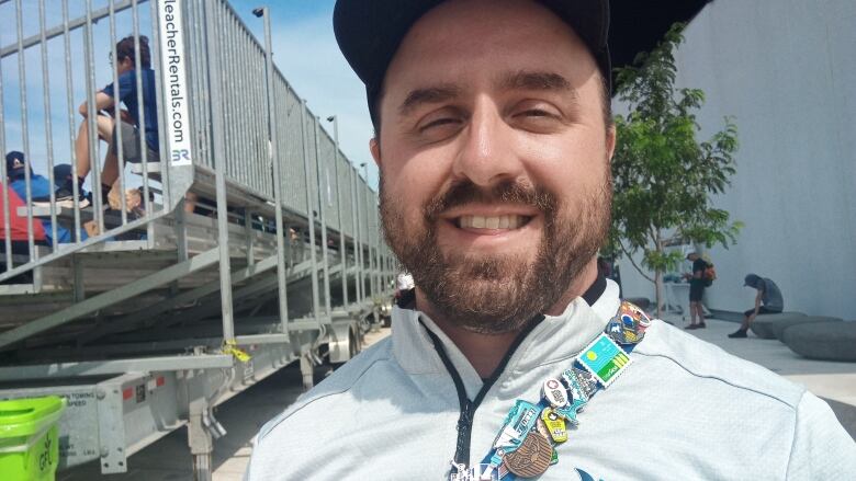 A man stands in front of some bleachers smiling. His bag strap has a lot of pins on it.