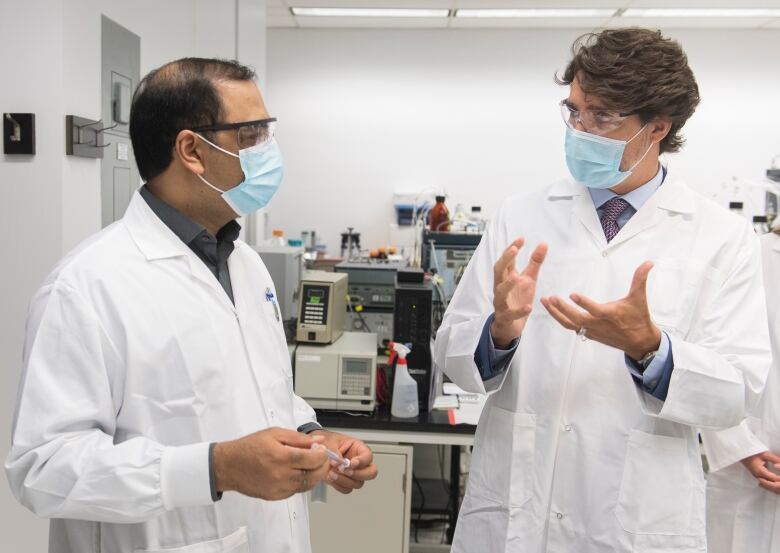 Two men wearing lab coats, face masks and safety glasses in conversation. One man holds a vaccine needle.