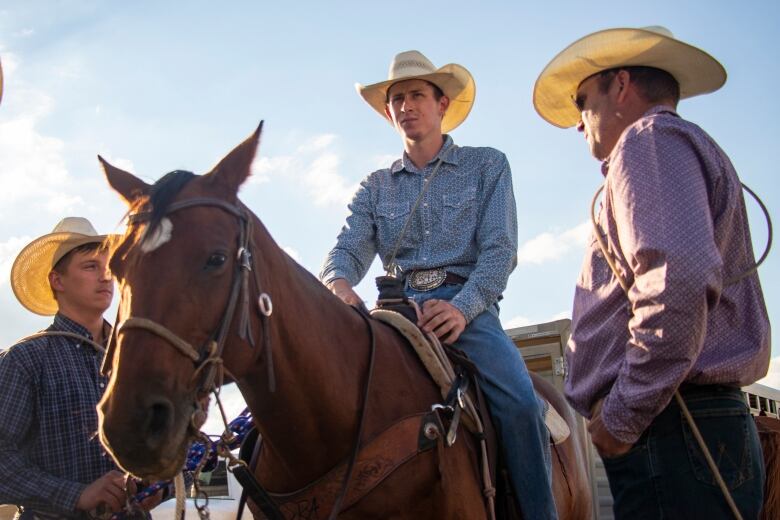 Two cowboys stand by a man sitting on a horse with the sun behind them.