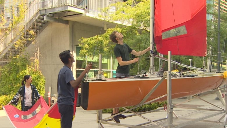 Two people work on a sailboat mounted on a table, with a third carrying a sail behind them.