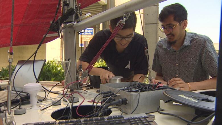 Two people work on equipment under a sailboat.