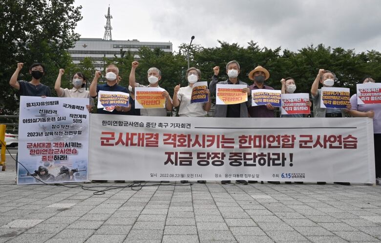 Several people in COVID-19 masks stand behind a banner with Korean writing, some holding their own signs.