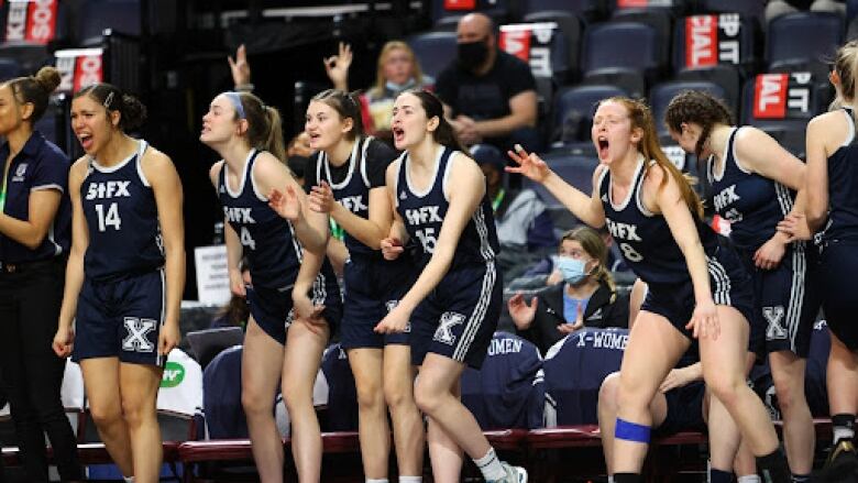 Women in basketball uniforms yell on the sidelines of a basketball court.