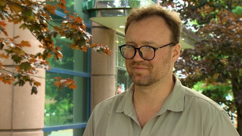 A man with glasses stands in front of a tree outside. 