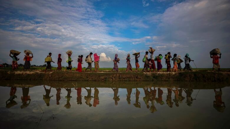 A line of people, some carrying bundles and baskets on their head, walk a long a river bank. 