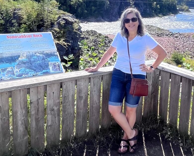 A person wearing sunglasses stands in front of a wooden rail at a lookout over a forest, a beach and a body of water.