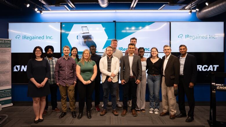 Fourteen people stand together in front of computer screens.