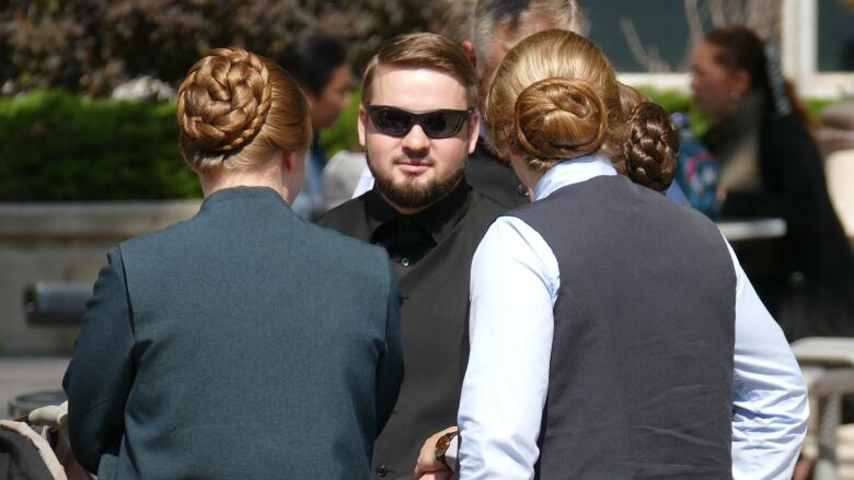 A man in sunglasses stands behind two women wearing buns in their hair and long, pleated skirts.