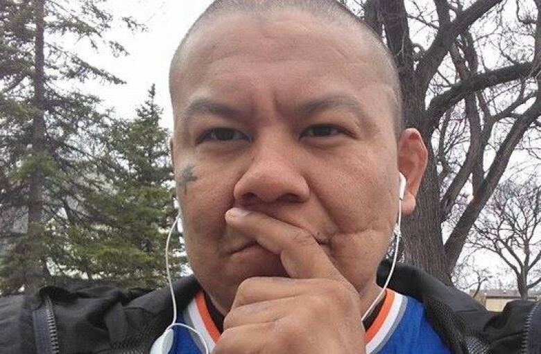 An Indigenous man is seen wearing a pair of earbuds. He looks quizzical, with his fingers on his mouth.