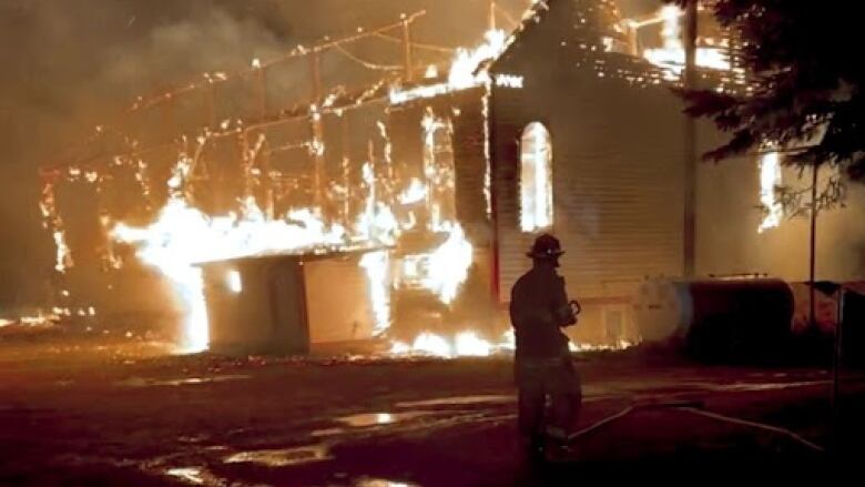 A firefighter stands in front of a burning structure. 