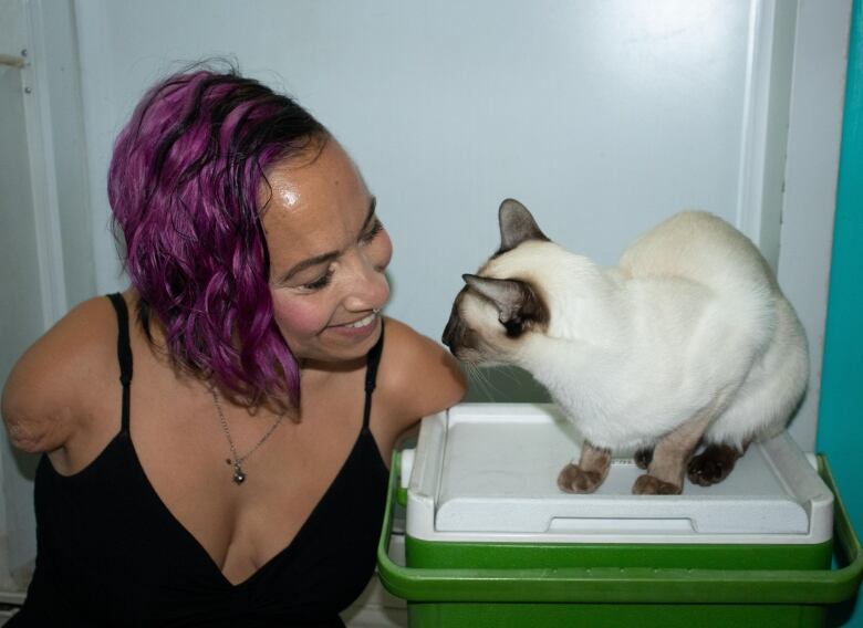 A smiling woman in a black dress, with choppy purple hair and no arms, stares into a white Siamese cats eyes lovingly. 