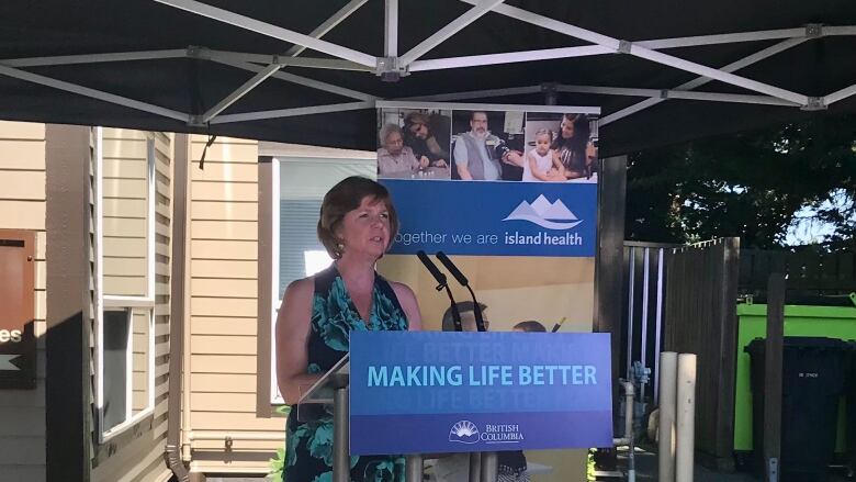 Sheila Malcolmson, a white woman wearing a black dress, stands at a podium with the words 'MAKING LIFE BETTER'.