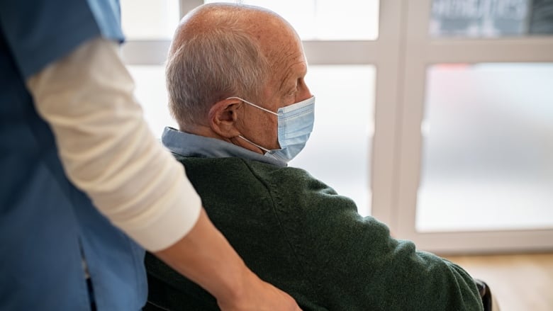 Nurse assists a sad elderly man using wheelchair wearing surgical face mask.