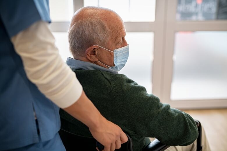 Nurse assists a sad elderly man using wheelchair wearing surgical face mask.