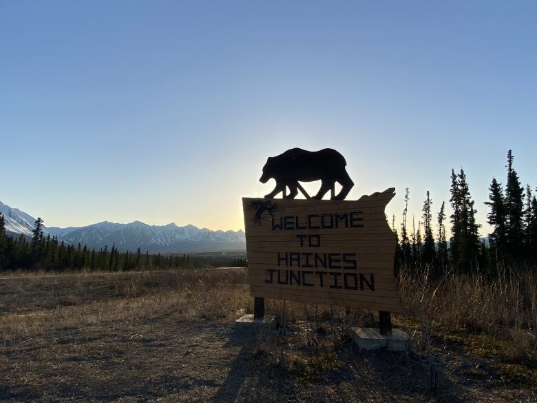 A silhouette of the Welcome to Haines Junction sign at sunrise with snowy mountains in the background.  