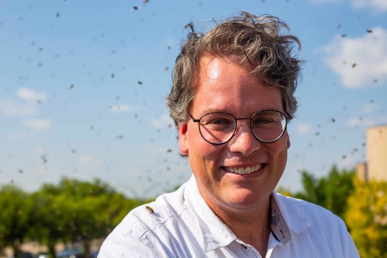 A smiling man in a white shoulder has a tiny bee on his shoulder as countless bees swarm around him.