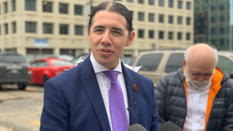 A man in a blue suit jacket and purple tie stands outside in a parking lot, in front of news media microphones.