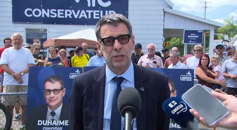 A man with thick rimmed glasses stands in front of a crowd of supporters. 