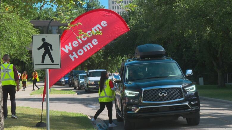 A woman in a green vest talks to a person in a black SUV outside the University of Calgary. there is a red flag that says welcome home off to the side. 