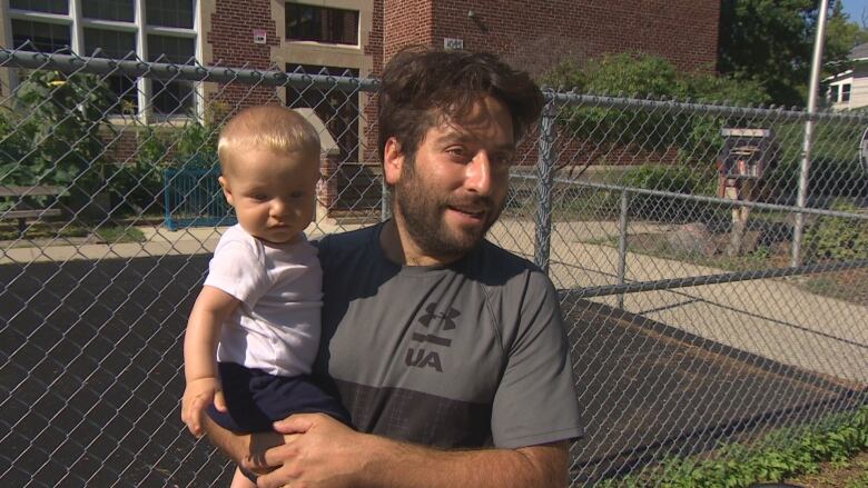 A man with a beard holds a baby as he talks outside of a school.