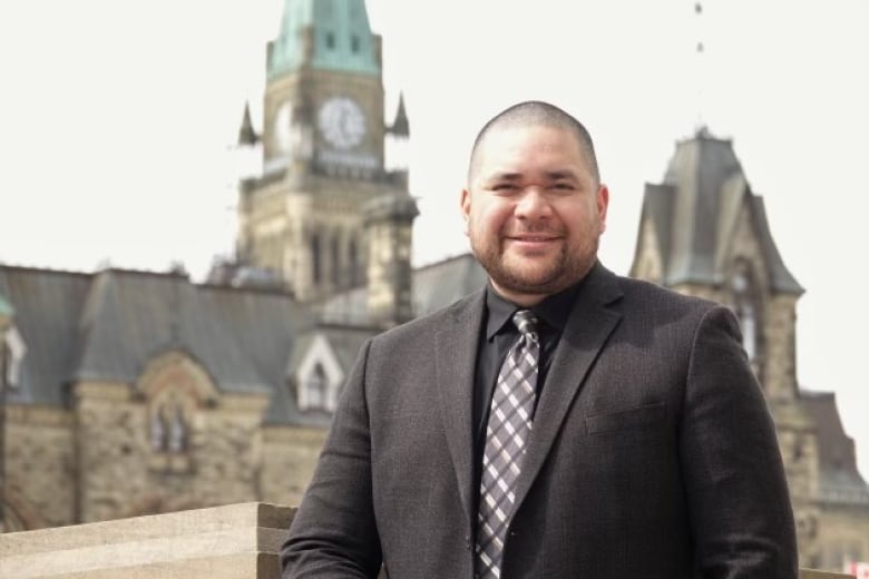 A man wearing a suit and patterned tie stands, smiling, in front of a building. 