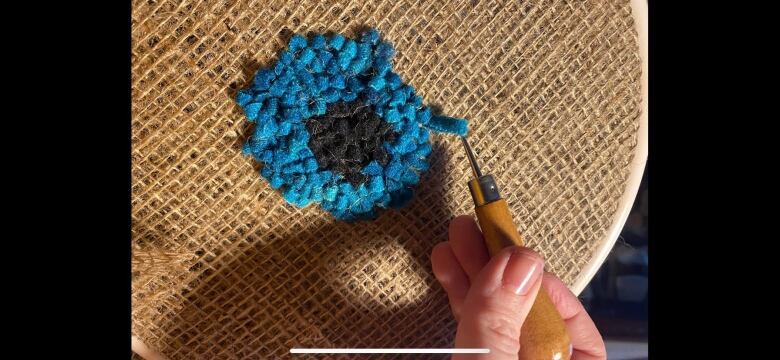 A bright blue flower is shown on burlap with a hand holding a hook.