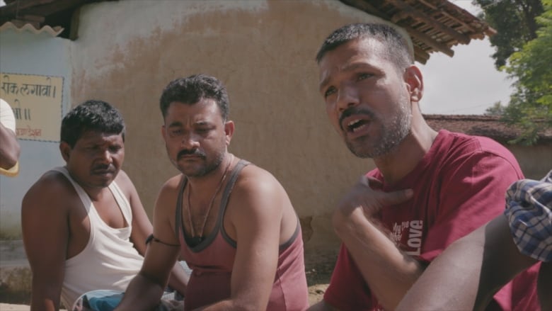 Three men sit next to one another. Behind them is a blank wall, and to their left a sign with writing in Sanskrit. 