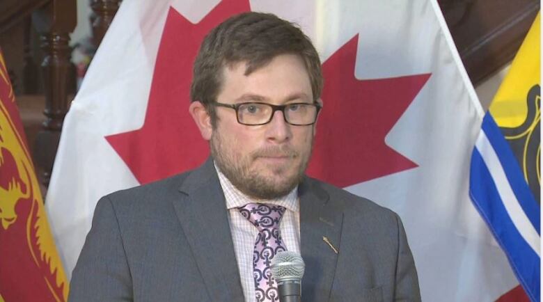 A man in a grey suit wearing glasses stands in front of the Canadian flag.