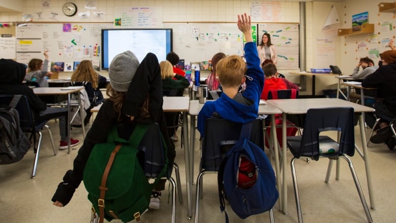 Students are seen interacting with their teacher and putting their hands up in a brightly lit classroom.