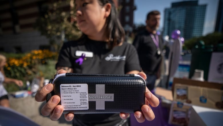 A woman with a purple ribbon on her chest holds out a kit with a cross and the word 'NALOXONE' on it.