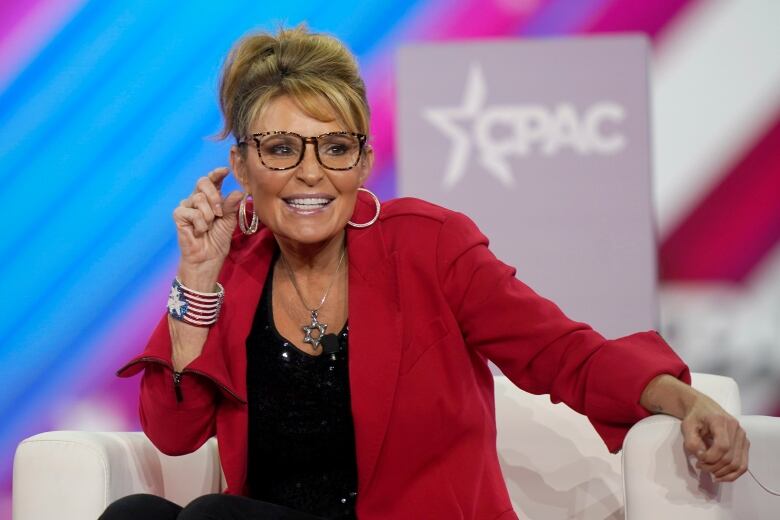 A smiling seated individual wearing glasses and a red blazer, with hair in an updo.