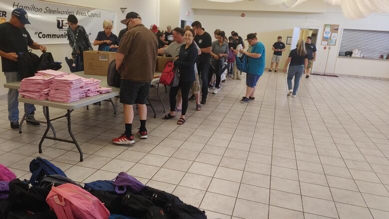 A large gathering of volunteers are filling two thousand backpacks with back-to-school supplies. Held at the Hamilton Steelworkers Area Council, the tables are lined with multicoloured bags, clothes and schoolwork supplies. 