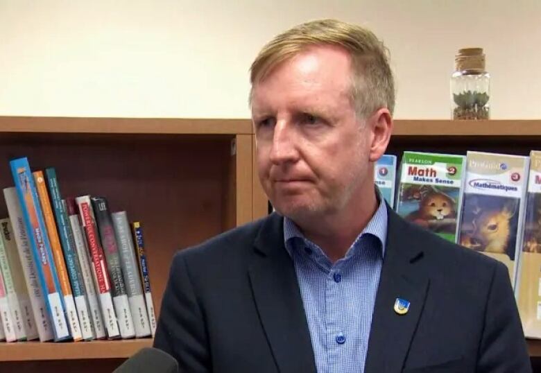 A man wearing a blue blazer and a blue unbuttoned shirt stands in front of a bookcase in a school classroom. 