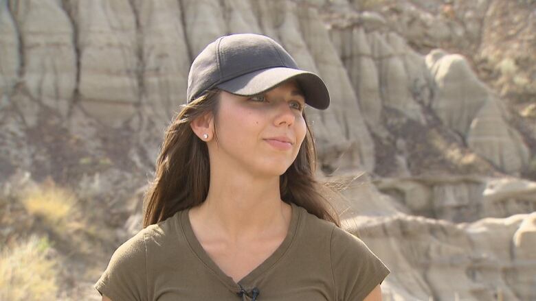 A woman in a green shirt wearing a black baseball cap stands in front of badlands rock. 